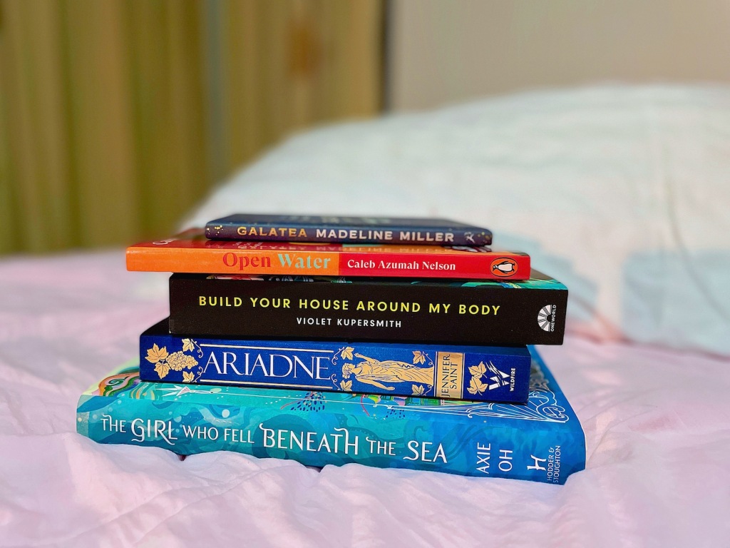 Photo shows a stack of books on a bed. From top to bottom: Galatea by Madeline Miller, Open Water by Caleb Azumah Nelson, Build Your House Around My Body by Violet Kupersmith, Ariadne by Jennifer Saint, and The Girl Who Fell Beneath the Sea by Axie Oh