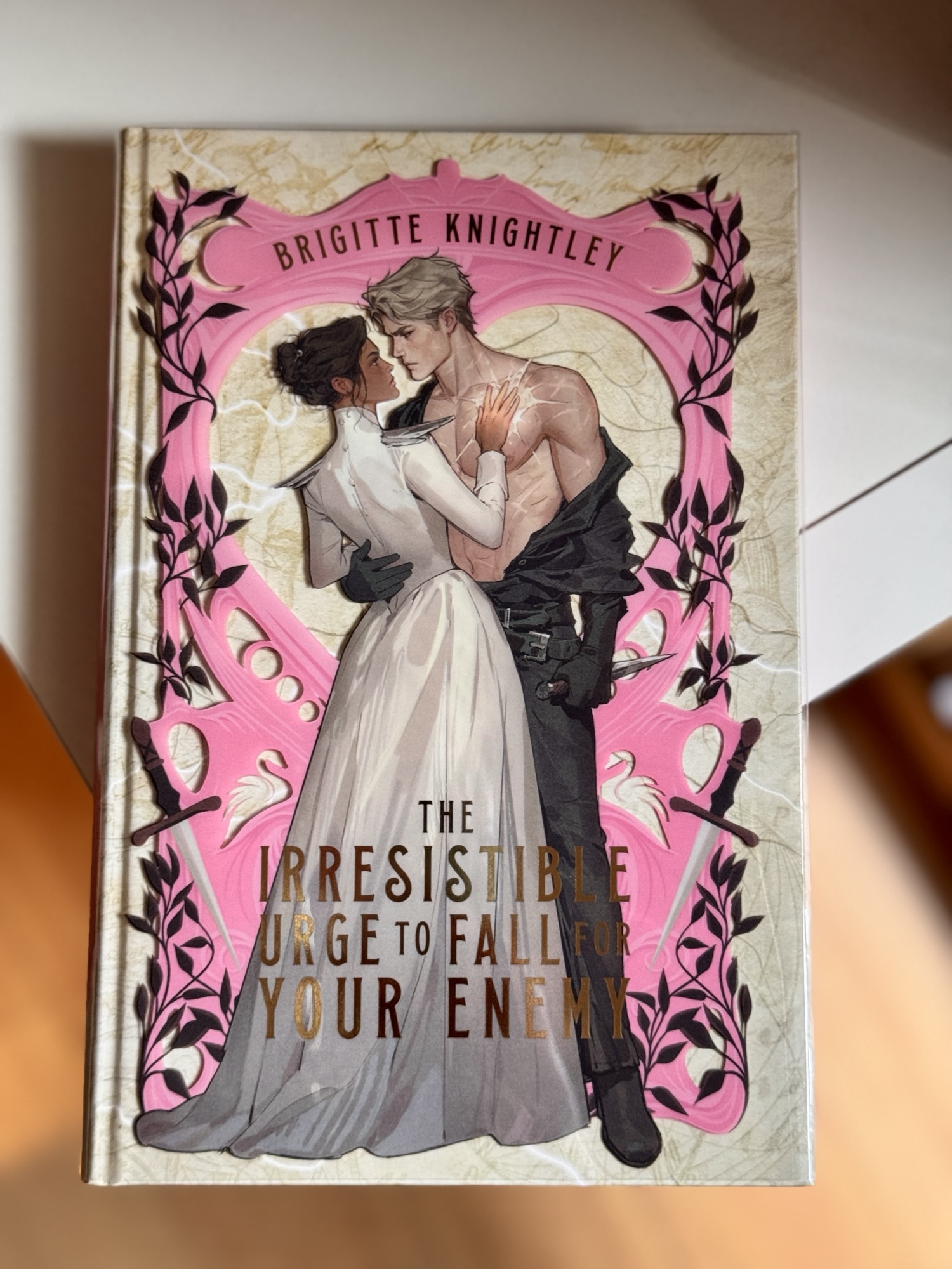 A flat lay image of the book The Irresistible Urge to Fall for Your Enemy by Brigette Knightley. The book has a white and pink background and the on the foreground are a fair-skinned half-naked man and a dark-skinned woman embracing.
